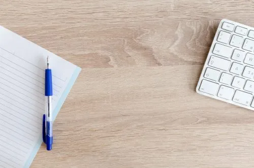 A pen on a lined notebook beside a keyboard on a wooden desk.