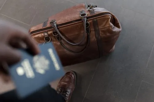 Hand holding a passport next to a brown leather duffel bag on the floor.