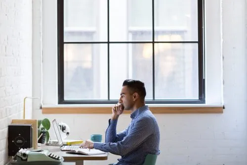Person working on a laptop at a desk near a large window.