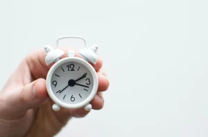 A hand holds a small white alarm clock showing 10:10.