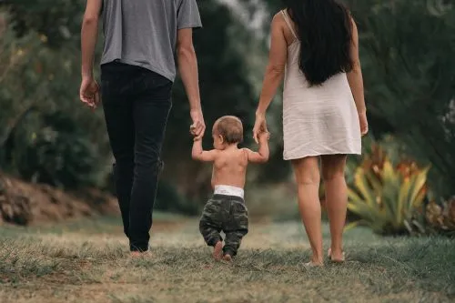 A toddler walks hand-in-hand with two adults on a grassy path.