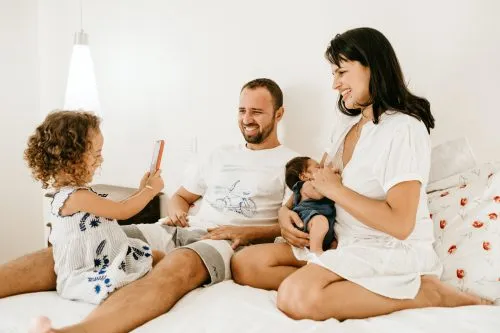 A family sits on a bed, smiling, with a young girl taking a photo and a woman holding a baby.