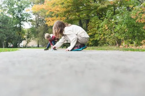 Two children crouching and drawing on a park path with autumn trees in the background.