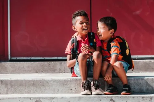 Two young boys with backpacks sit on steps laughing together.