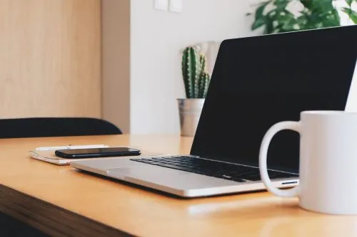 Open laptop, smartphone, and coffee mug on a wooden desk with a potted cactus plant.