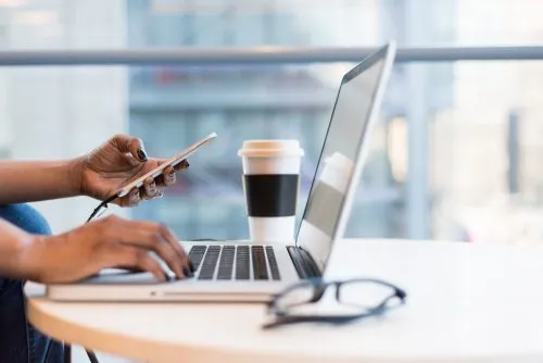Person using a laptop and smartphone at a table with a coffee cup.