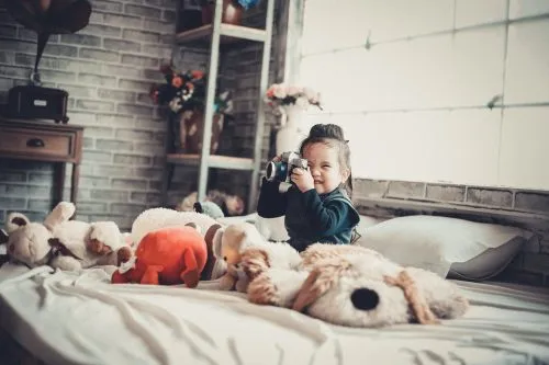 Child sitting on a bed with stuffed animals, playfully using a camera.