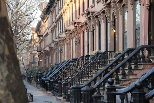 A row of brownstone buildings with ornate stoops on a tree-lined street.