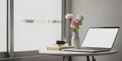 A laptop, coffee cup, book, and vase with flowers on a round table by a window.