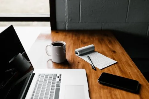 Laptop, coffee mug, notepad, pen, and smartphone on a wooden desk by a window.
