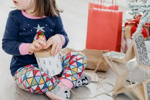 Child in colorful pajamas opening a gift with holiday decorations and a red bag nearby.