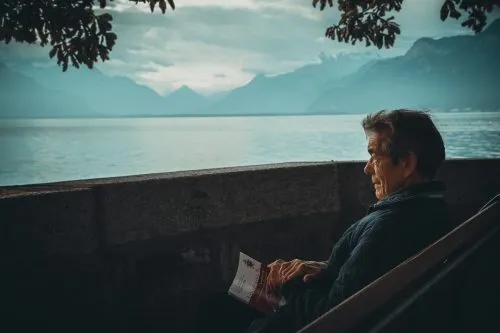 Older person sitting on a bench by a lake, holding a paper, with mountains in the background.