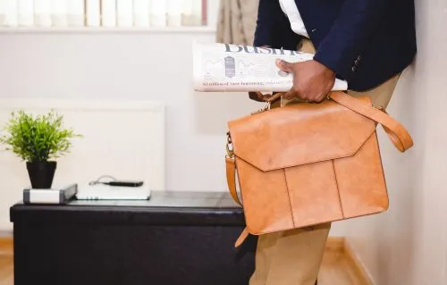 Person holding a leather bag and newspaper, standing by a table with a plant and notebook.