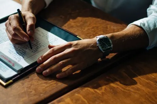 Person writing in a notebook on a wooden table, wearing a watch.