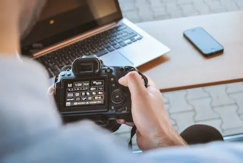 Person holding a DSLR camera with a laptop and smartphone on a table.