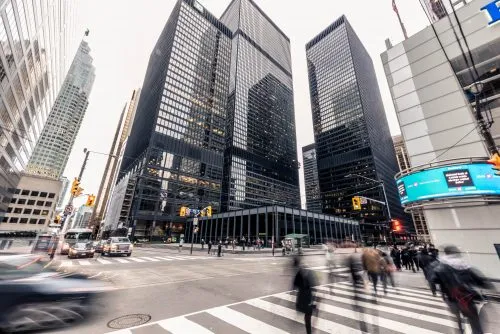People crossing a busy city intersection surrounded by tall skyscrapers.