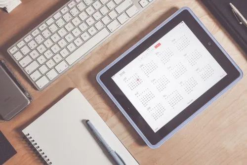 Tablet displaying a calendar placed on a wooden desk with a keyboard, notebook, and pen.