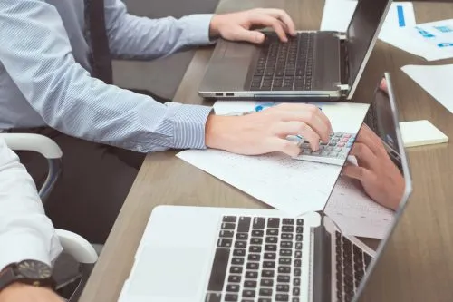 Two people working at laptops and using a calculator on a desk.