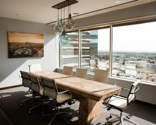 Sunlit conference room with a wooden table, modern chairs, and a cityscape view through large windows.
