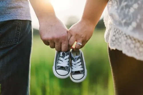 Couple holding baby shoes in a grassy field with sunlight.