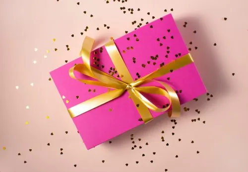 Pink gift box with a gold ribbon on a pink surface scattered with gold confetti.
