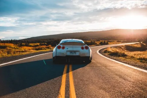A white sports car drives down a winding rural road at sunset.
