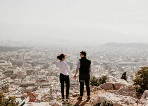 Two people stand on a rocky hill overlooking a sprawling cityscape.