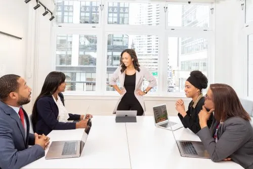 A businesswoman stands confidently at a table, leading a meeting with colleagues.