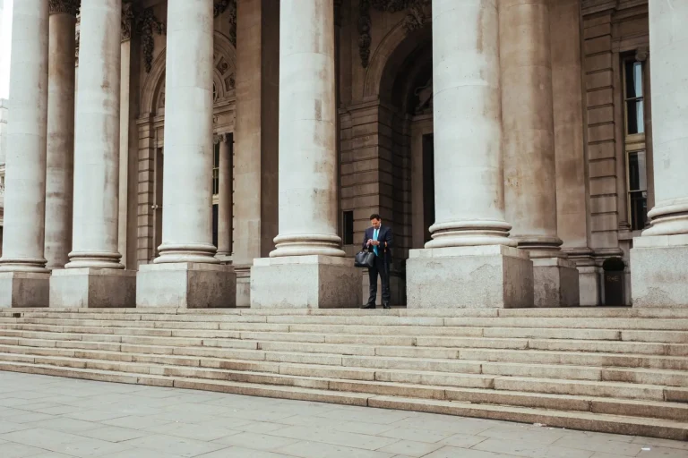 A person stands with a bag on the steps of a large building with tall stone columns.