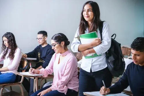 A student stands holding books in a classroom while others sit and write.
