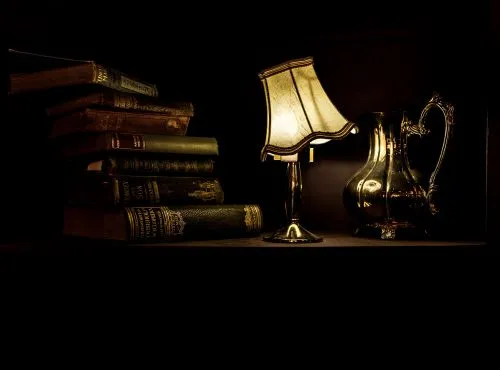 Stack of old books and a lit lamp beside a decorative pitcher on a dark shelf.