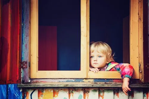 A young child in a colorful jacket looks out of a painted wooden window.