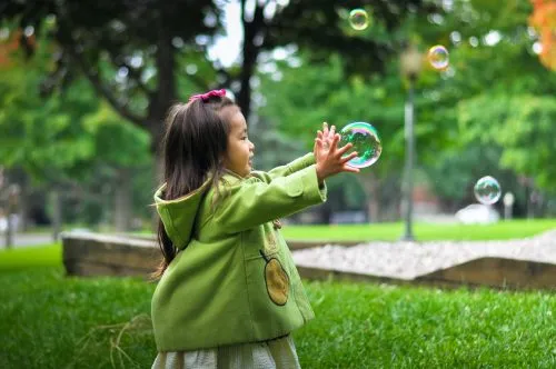 A child in a green coat plays with bubbles in a grassy park.