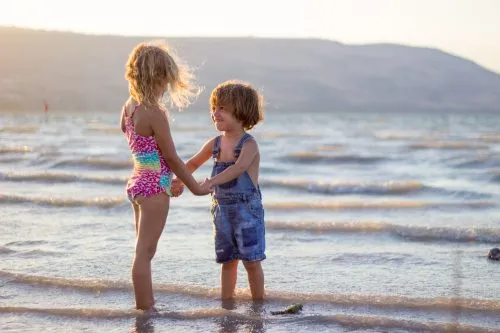 Two children holding hands, standing in shallow beach water at sunset.