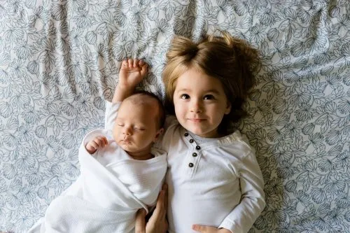 Two children lie on a patterned blanket, a newborn swaddled and a toddler in a white shirt.