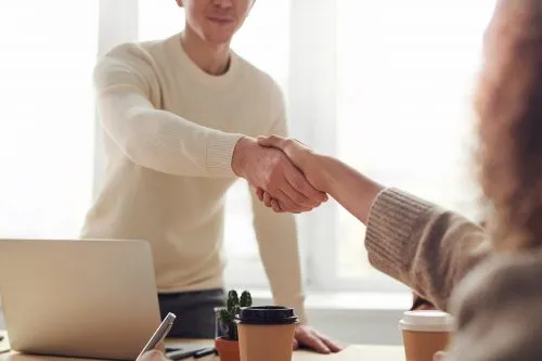 Two people shaking hands over a desk with coffee cups and a laptop.
