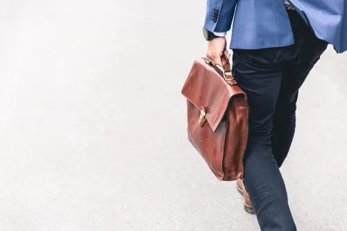 Person walking with a brown leather briefcase on a gray pavement.