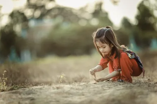 A young girl in a red dress crouches and plays with soil in a grassy field.