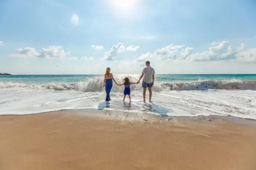 A family holds hands facing the ocean on a sunny beach.