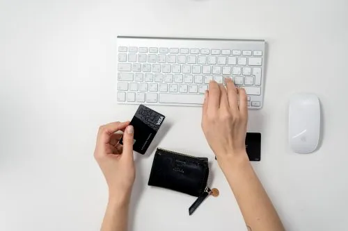 Hands holding a credit card near a keyboard and a black wallet on a white desk.