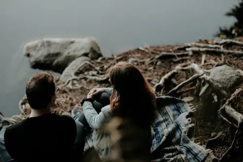 A man and woman sit on a blanket by a lakeshore, surrounded by rocks and roots.