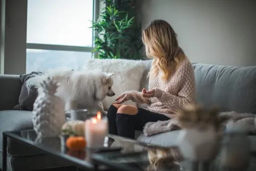 Woman sitting on a sofa, petting a small white dog, with candles on a glass coffee table.