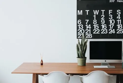 Minimalist workspace with a wooden desk, computer, plant, and wall calendar.