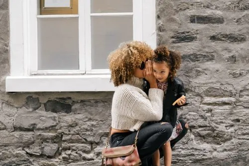 Woman whispering to a young girl outside a stone building.