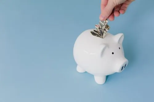 Hand placing dollar bills into a white piggy bank on a blue background.