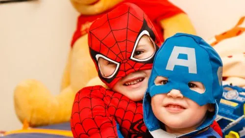 Two children in Spider-Man and Captain America costumes smile together.