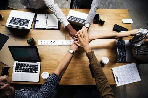 A group of people seated at a table join hands over laptops and documents.
