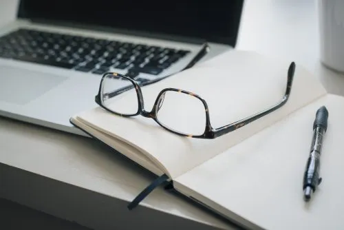 Glasses and a pen on an open notebook beside a laptop.
