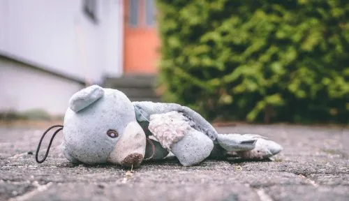 Worn teddy bear lying on a paved ground outdoors near a house and greenery.