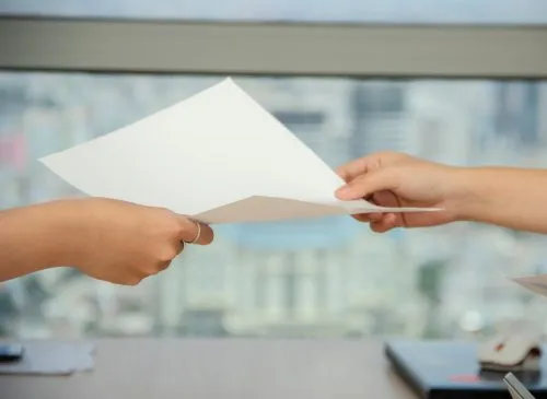 Two hands exchange a sheet of paper in front of a blurred cityscape background.